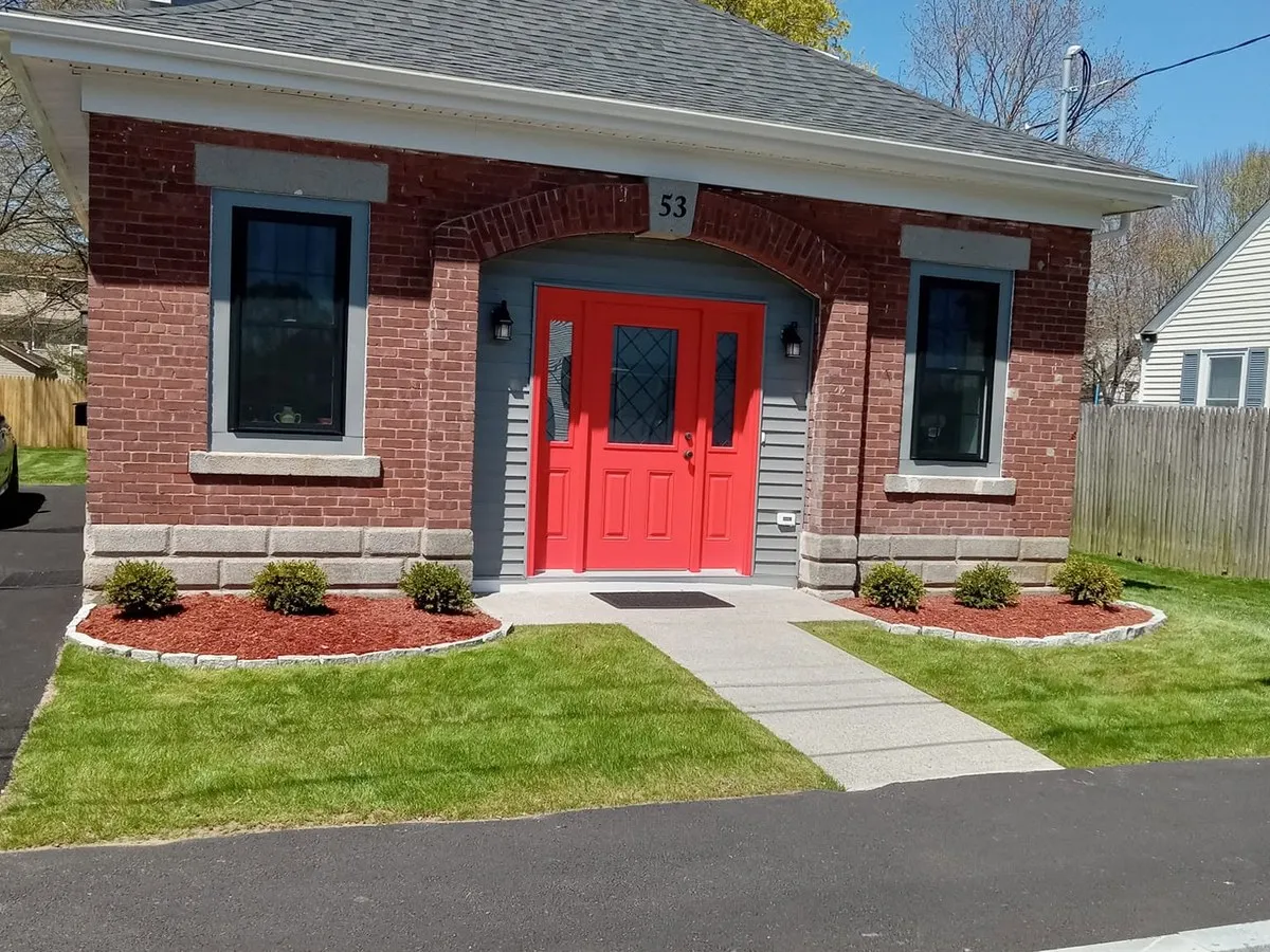 Red mulch beds with granite cobblestone edging and boxwood shrubs flanking the walkway of a brick house with a red door
